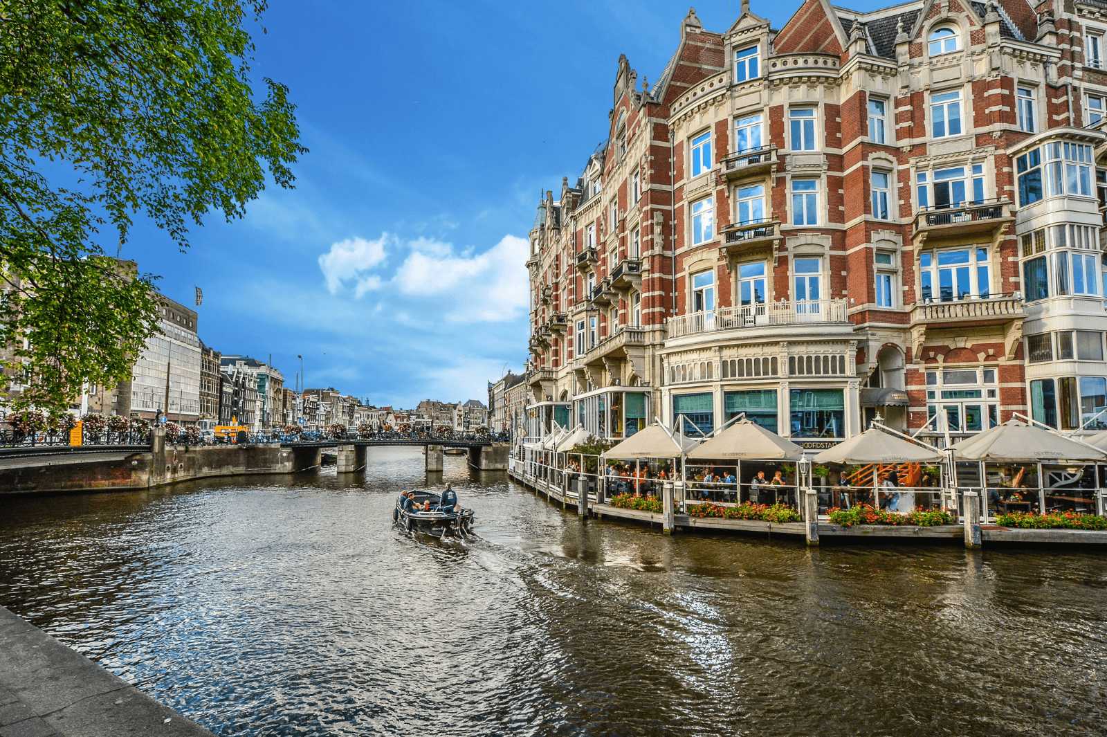 Uitzicht op een schilderachtige gracht in Amsterdam met een bootje op het water, een historische brug op de achtergrond en een statig gebouw met terrassen aan het water onder een heldere blauwe lucht.