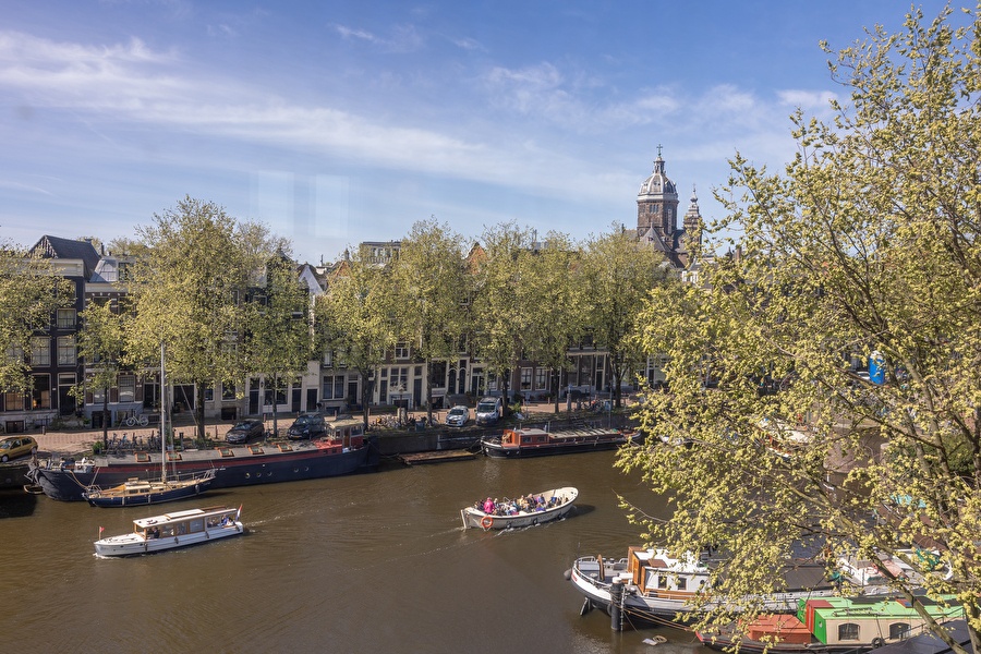 View of the Amsterdam canal with houseboats, trees, and the Basilica of Saint Nicholas in the background.