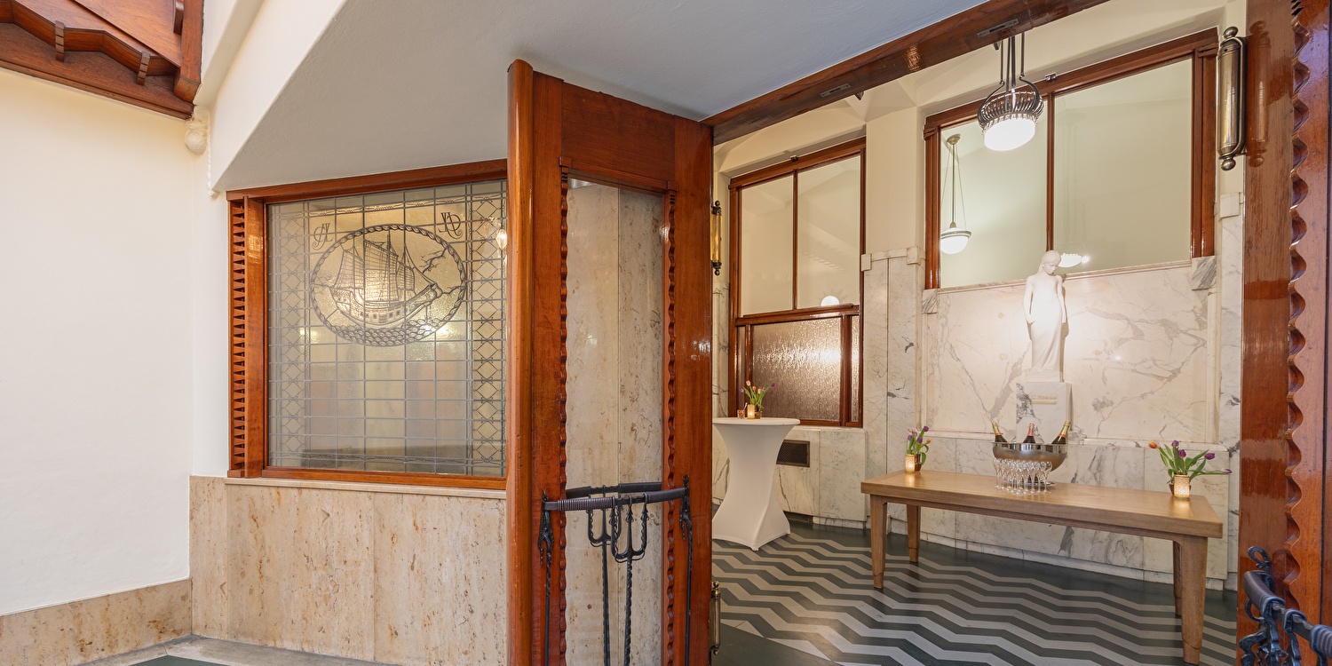 Detail van de Van der Meij hal in Grand Hotel Amrâth Amsterdam met marmeren wanden, glas-in-loodraam met scheepsmotief, houten accenten en zigzag-patroon op de vloer. Sfeervolle ruimte voor fotoshoots en evenementen.