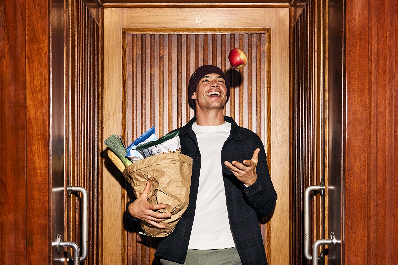 Man in de historische Paternosterlift van Grand Hotel Amrâth Amsterdam, met boodschappen en een appel die hij de lucht in gooit, tijdens een MR MARVIS fotoshoot.