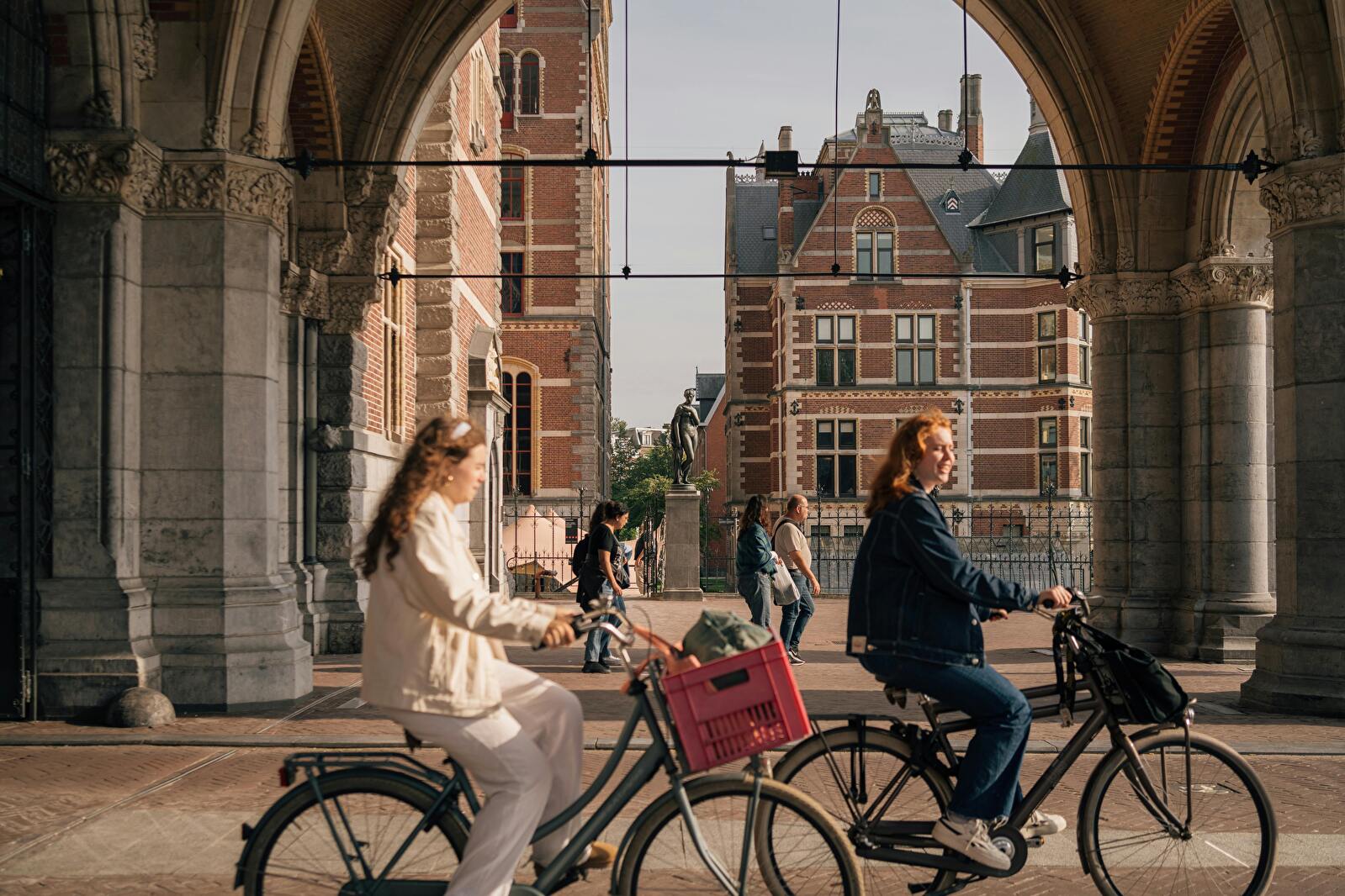 Twee vrouwen fietsen onder de doorgang van het Rijksmuseum in Amsterdam, met het karakteristieke bakstenen gebouw en een standbeeld op de achtergrond.