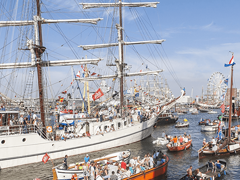 Drukke en feestelijke taferelen op het IJ tijdens SAIL Amsterdam, met tall ships, historische boten en veel mensen onder een stralende blauwe lucht.