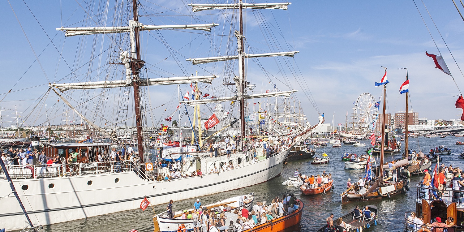 Drukke en feestelijke taferelen op het IJ tijdens SAIL Amsterdam, met tall ships, historische boten en veel mensen onder een stralende blauwe lucht.