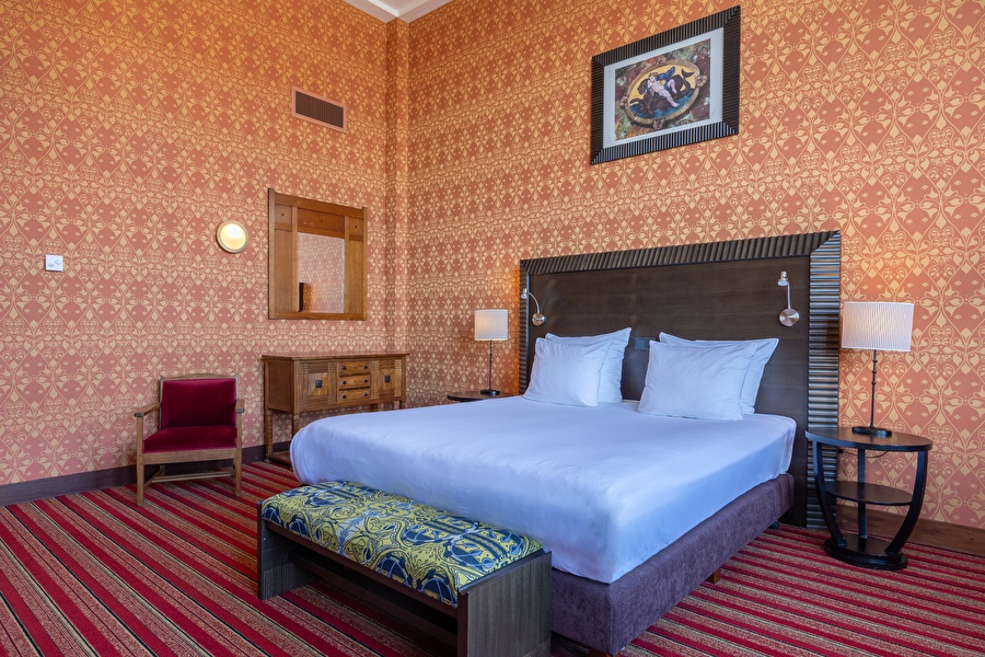 Spacious Superior Deluxe room with designer wallpaper, comfortable armchair, and wooden chest of drawers.